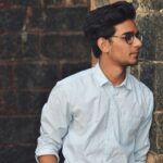 A stylish young man in glasses leans against a brick wall wearing a casual light blue shirt.